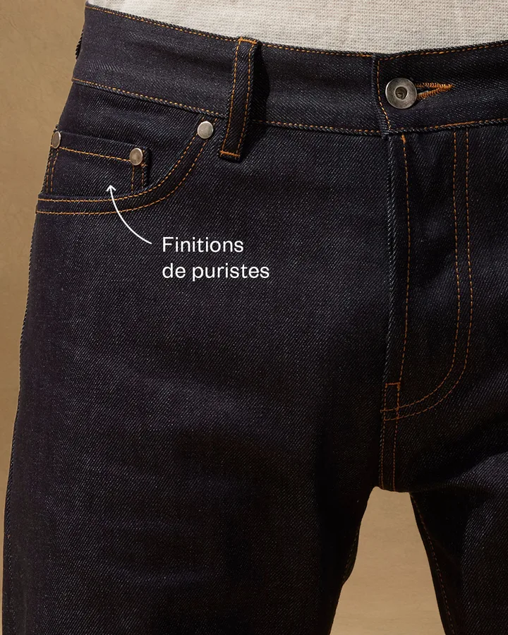 Gros plan sur les surpiqûres et la braguette boutonnée du denim.